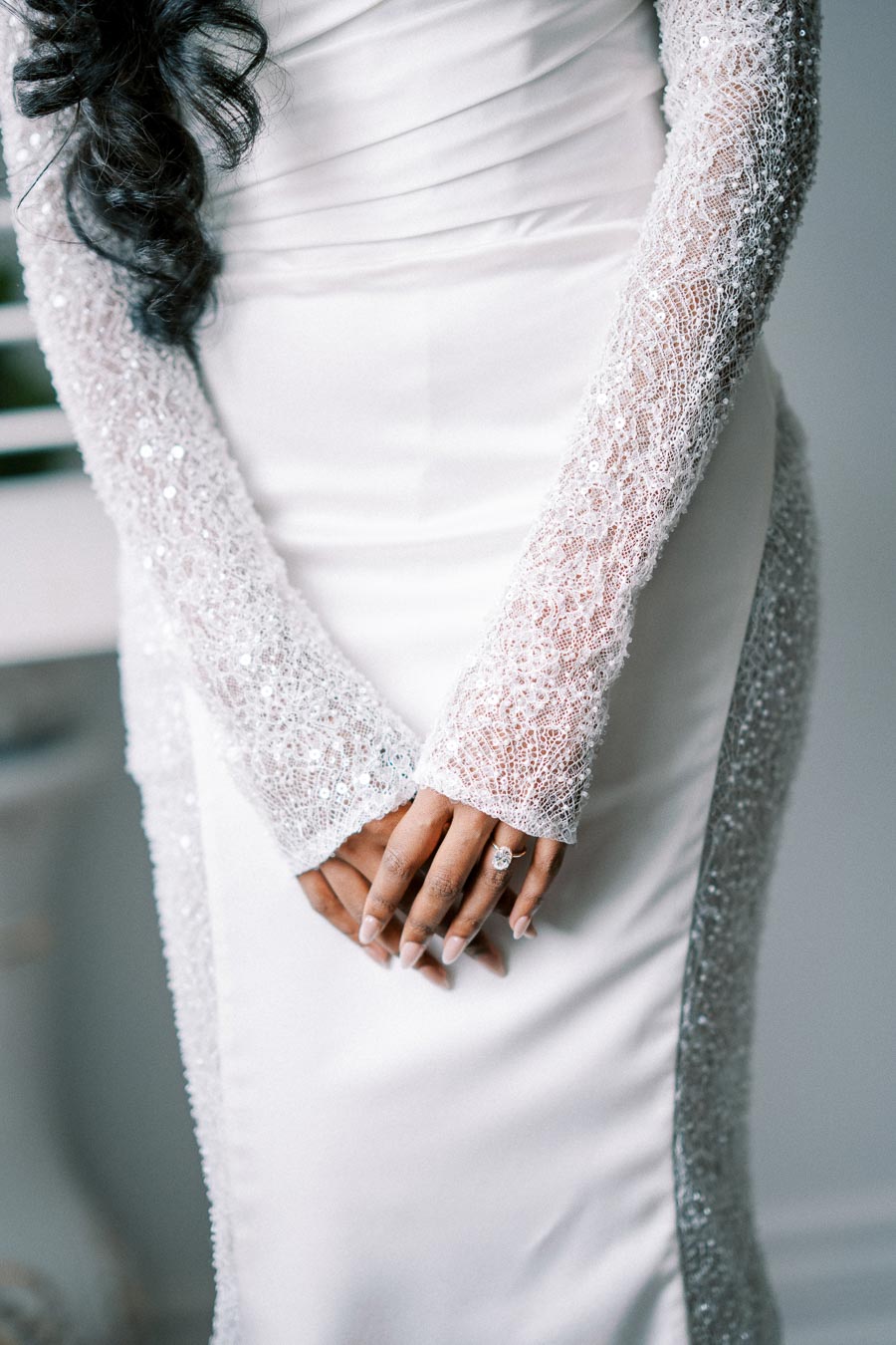Elegant bride in a stunning white wedding dress with lace sleeves, hands clasped with a sparkling engagement ring.