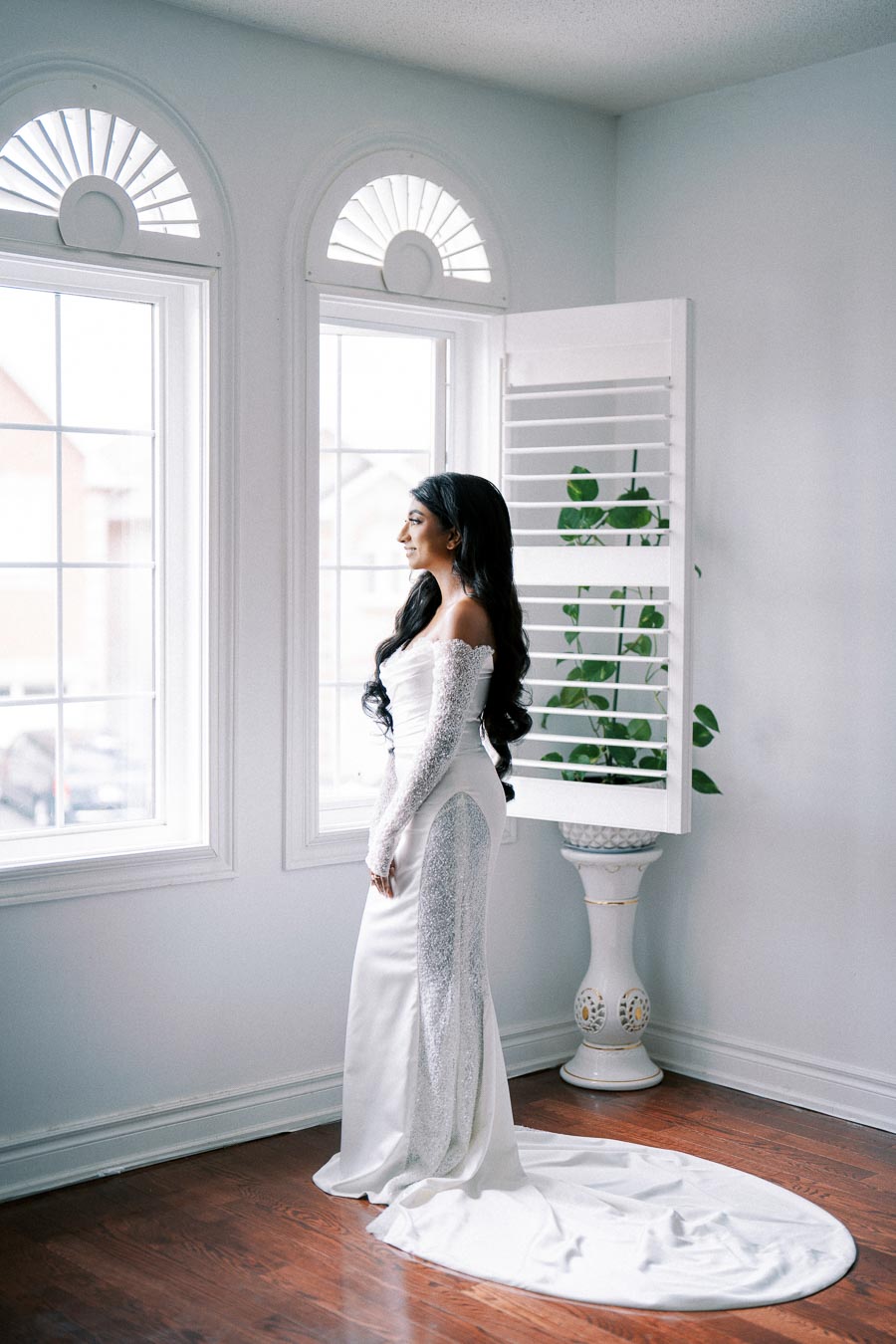 Bride standing in elegant white wedding dress with long train, gazing out bright window in a serene room.