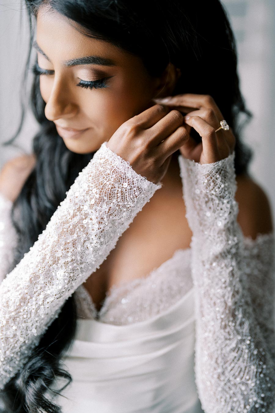 Elegant bride adjusting her earring, wearing a white lace wedding dress with intricate beadwork, capturing a serene moment before the ceremony.