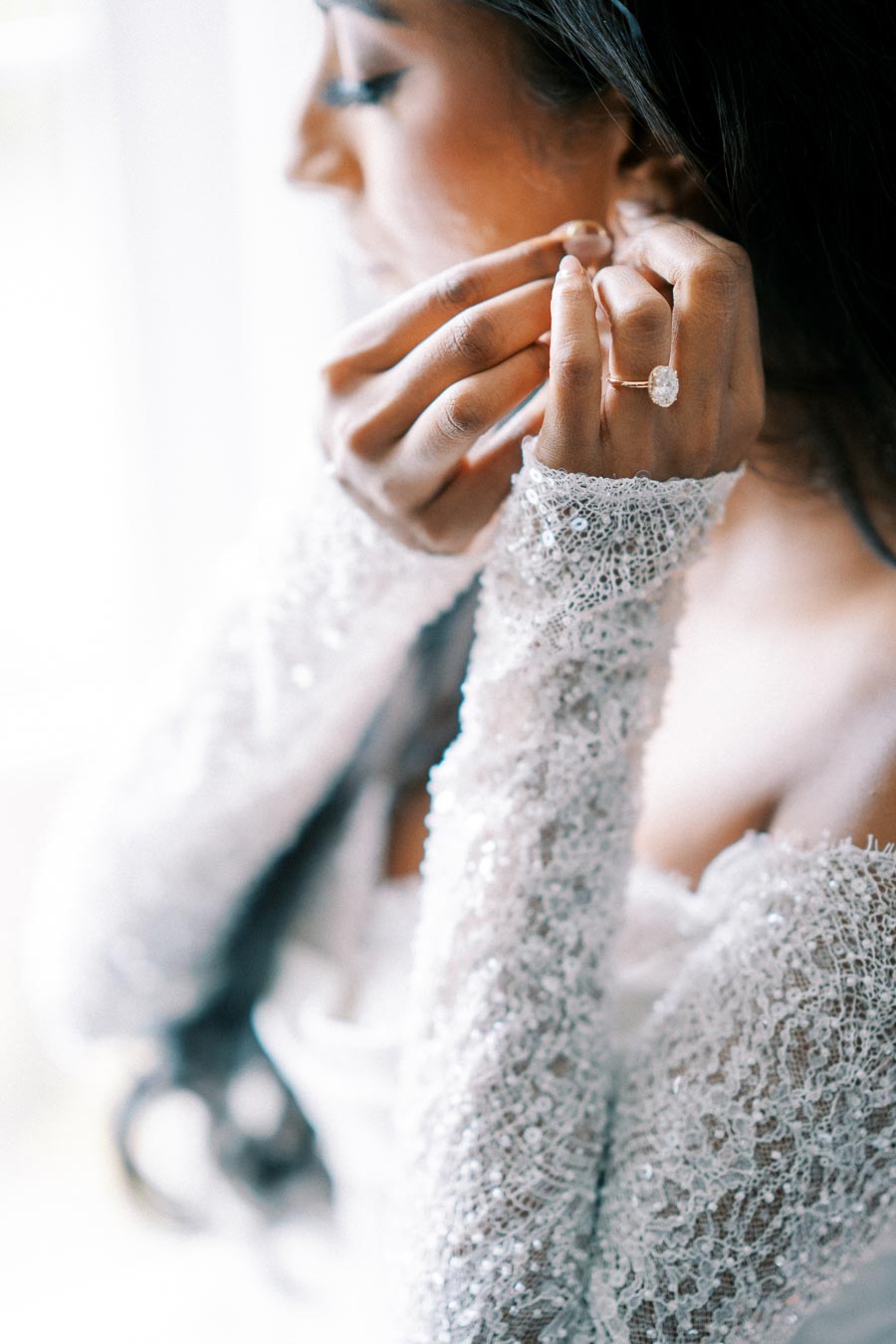 A bride in an elegant lace wedding dress adjusts her earring, showcasing a sparkling oval diamond engagement ring.