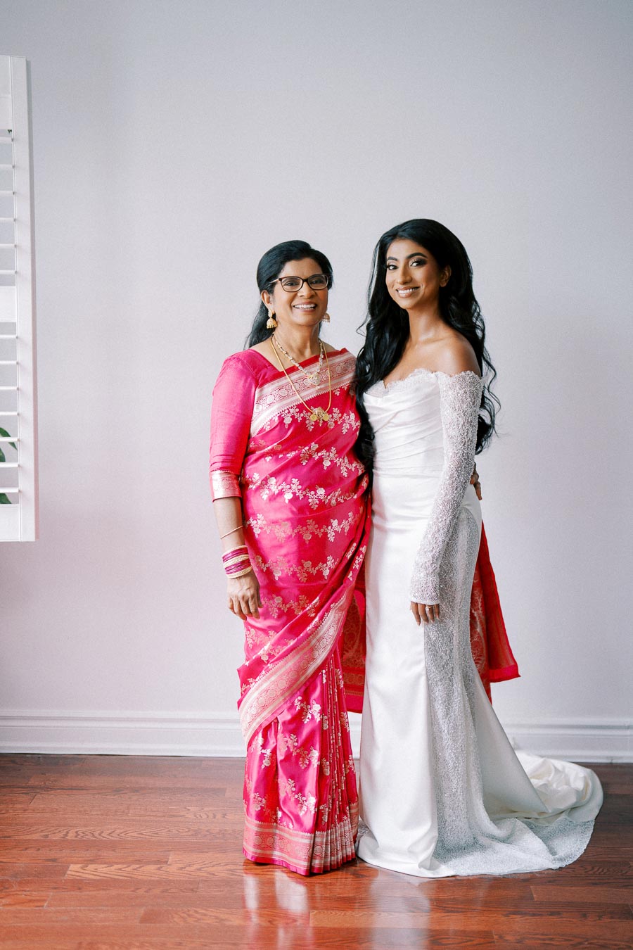 Two women smiling at a camera; one is wearing a vibrant pink saree adorned with gold accents, while the other is in an elegant white off-the-shoulder bridal gown.