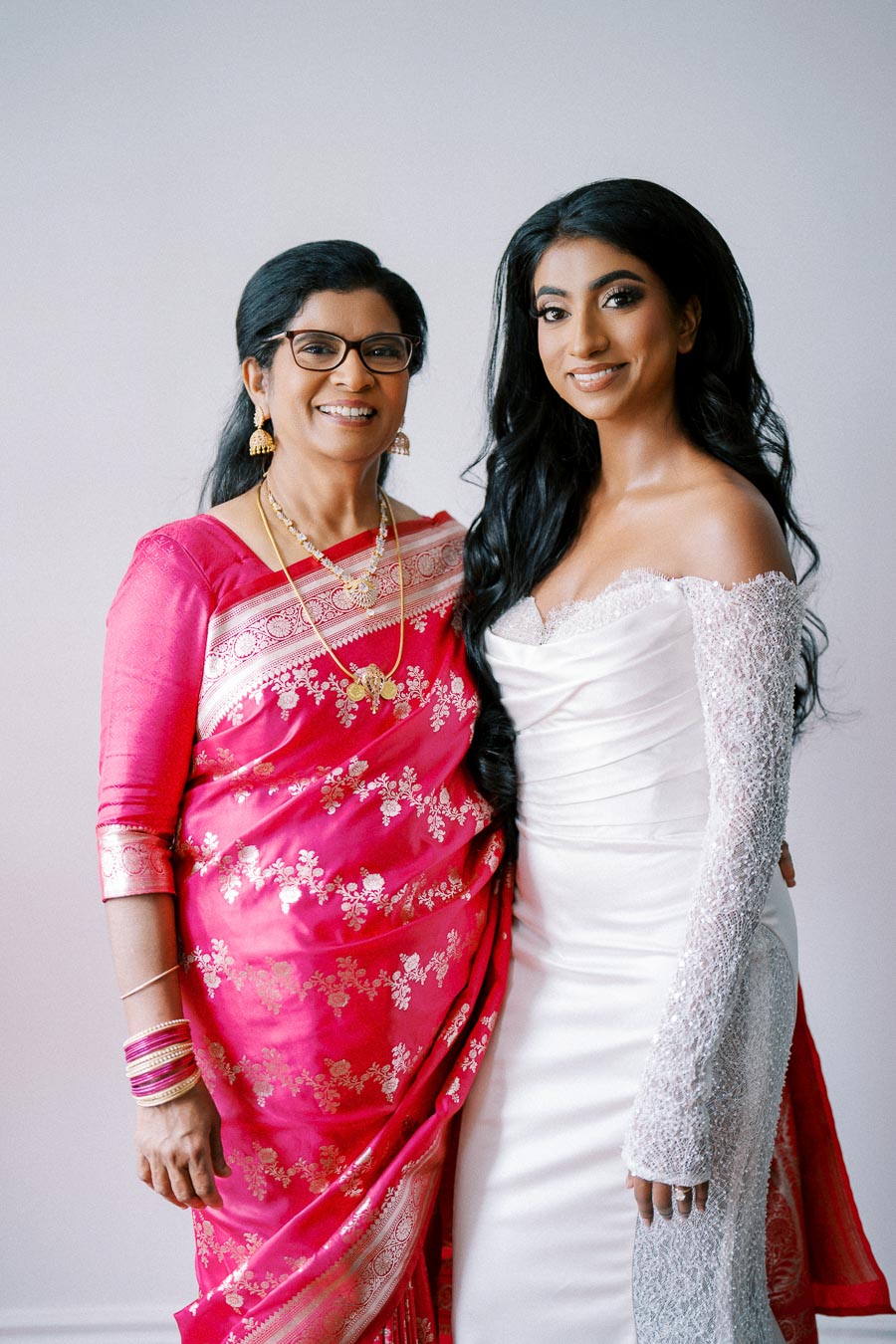 A bride in a white gown and a woman in a vibrant pink saree smiling together.