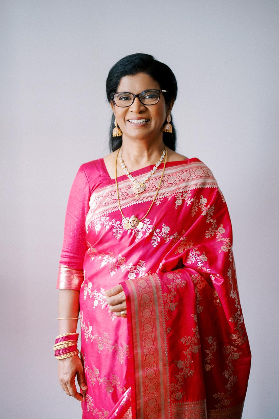 A woman in a vibrant pink and gold traditional saree, smiling confidently while wearing glasses, earrings, and necklaces against a plain background.
