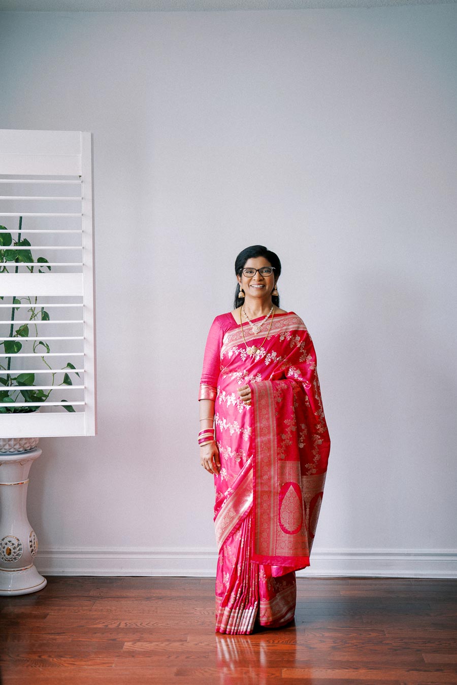 A woman in a vibrant pink saree stands indoors against a light-colored wall, smiling and displaying traditional attire, with a leafy plant beside a window in the background.