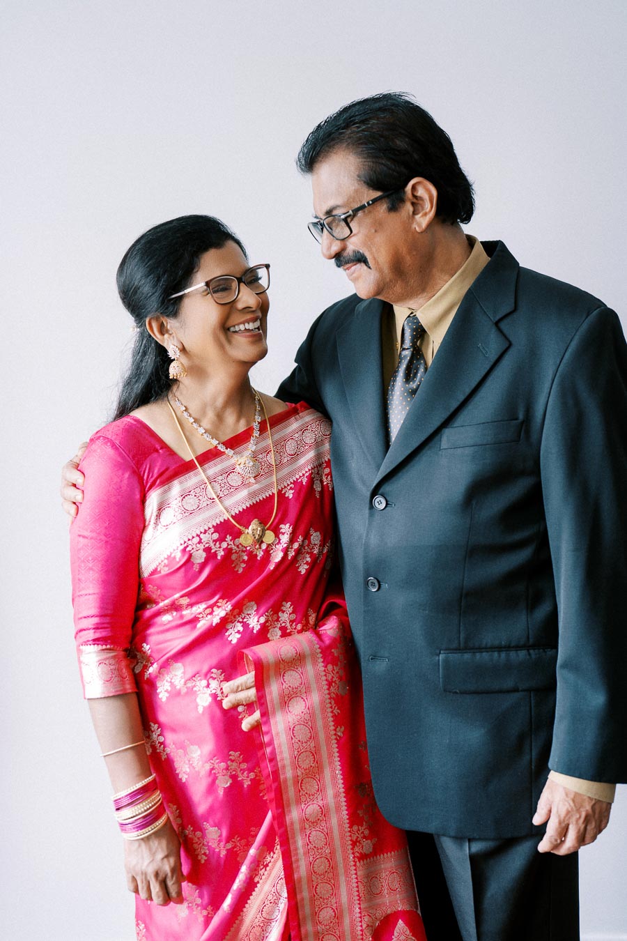 Elderly couple dressed elegantly, the woman in a pink sari with gold jewelry and the man in a dark suit, sharing a joyful moment and looking at each other.