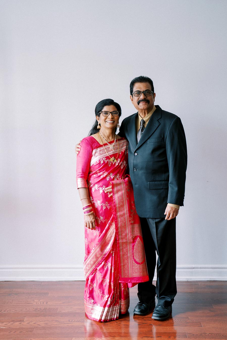 Elderly couple posing for a formal portrait, with the woman in a vibrant pink sari and the man in a black suit, standing on a polished wooden floor against a plain white wall.