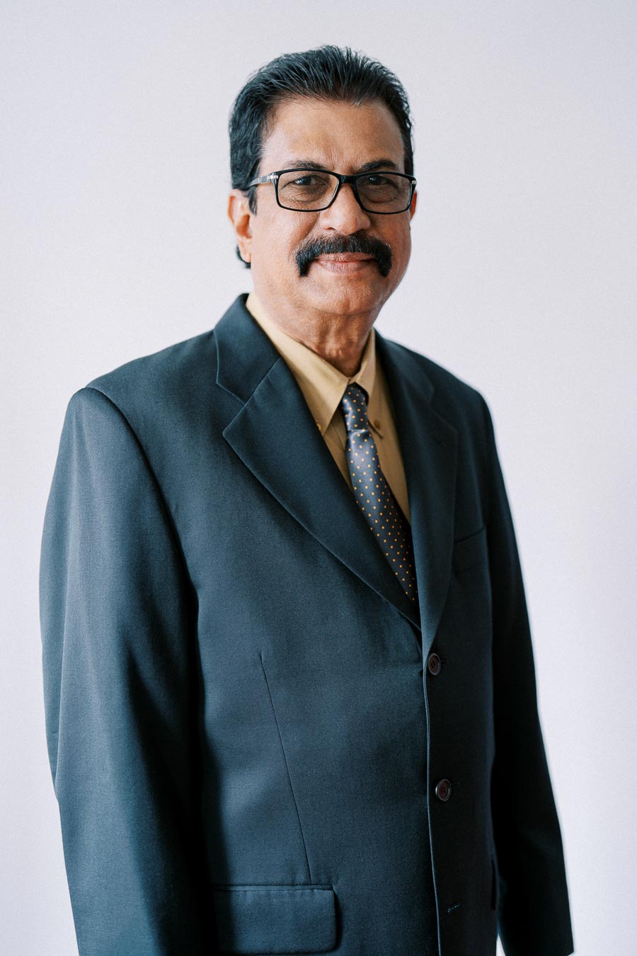 Portrait of a man with a mustache wearing glasses and a dark suit with a polka dot tie, standing against a plain background.