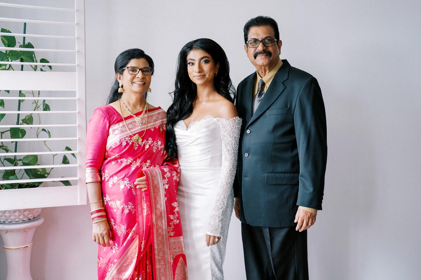 A bride in a white wedding gown stands between two older adults. The woman on the left is wearing a pink saree, and the man on the right is in a dark suit. They are posing indoors with a white wall and window blinds in the background.