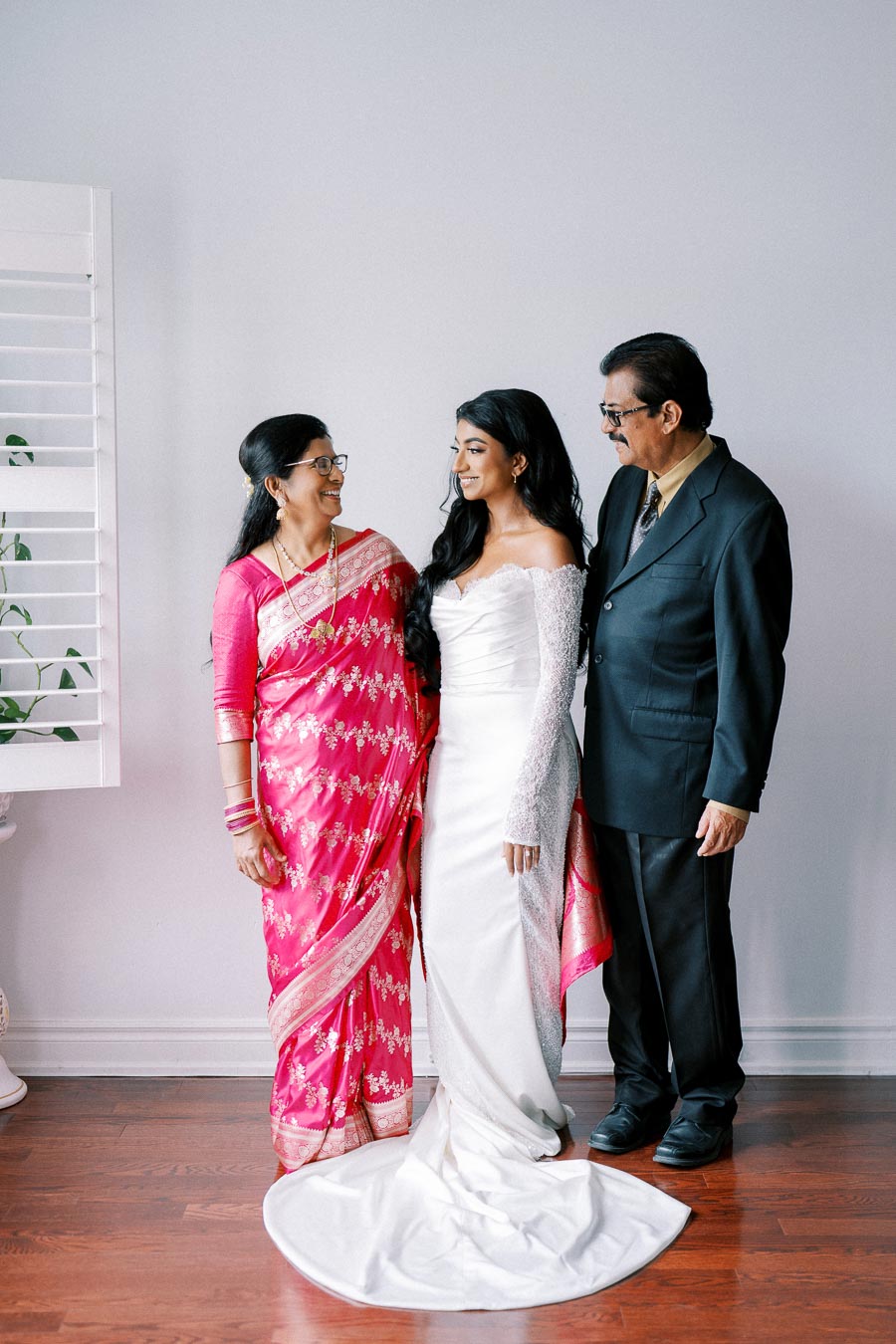 A bride in a white gown stands between two elegantly dressed parents; her mother wears a vibrant pink sari, and her father is in a dark suit. They are smiling at each other against a neutral indoor background.