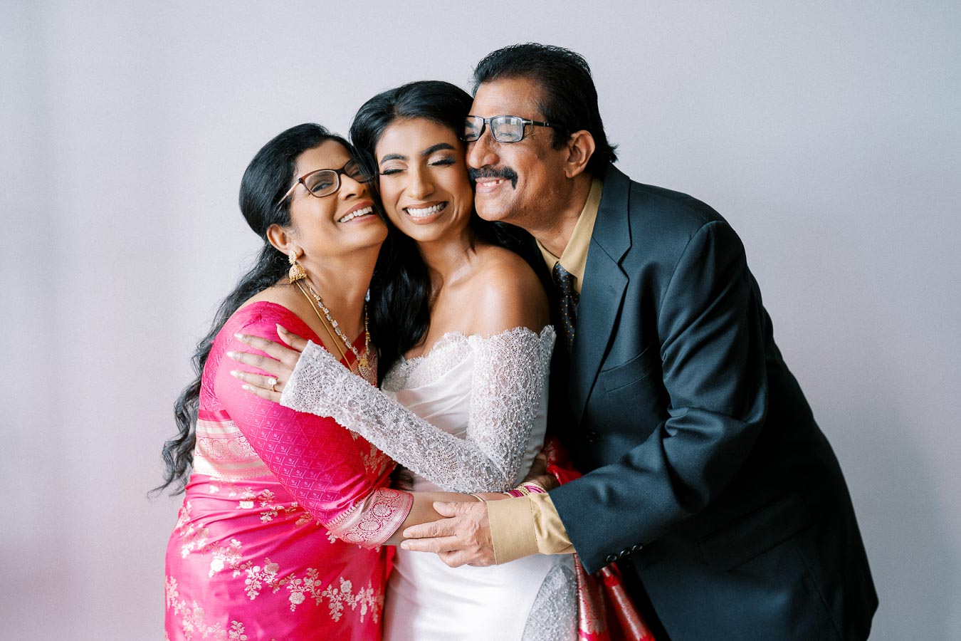 A bride in a white dress joyfully hugs her parents as they smile warmly, with her mother in a vibrant pink saree and her father in a black suit, against a neutral background.