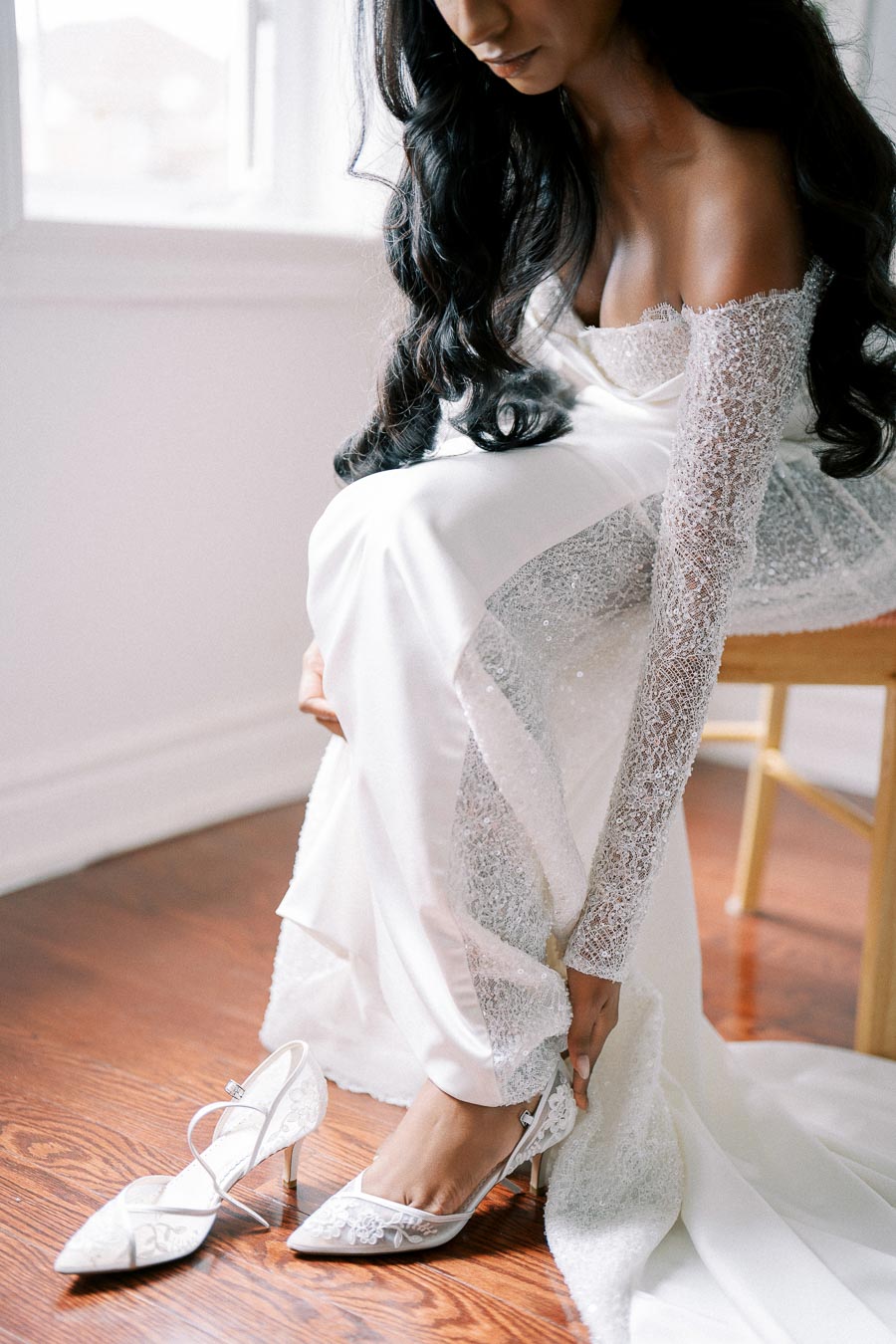 Bride in elegant lace wedding dress adjusting her white heels, sitting on a wooden floor in a softly lit room, preparing for her special day.