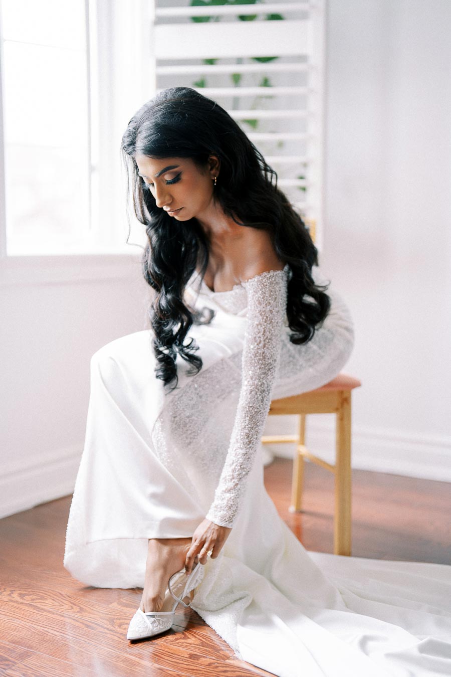 A bride in an elegant white wedding dress adjusts her shoe while sitting on a wooden chair near a window, showcasing her long, wavy hair and radiant complexion in natural light.