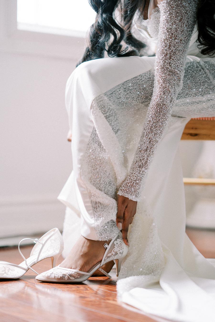 Elegant bride in a lace wedding gown putting on delicate white floral embroidery heels, captured indoors on a wooden floor.