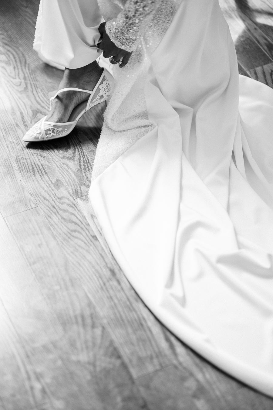Black and white image of a bride adjusting her elegant lace high-heeled shoe, with a flowing white wedding dress on a wooden floor.