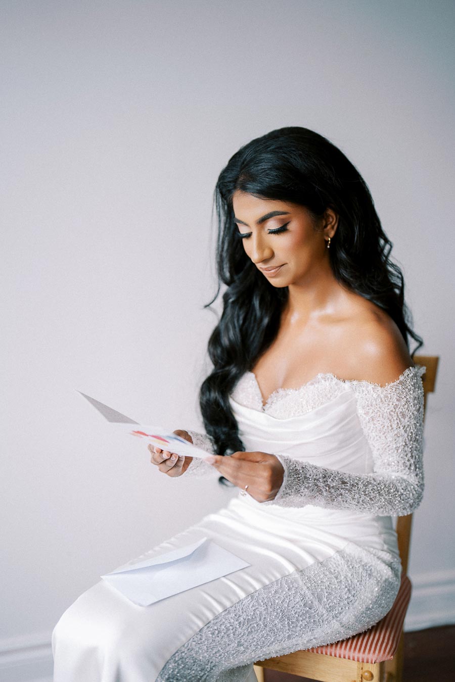 Bride with long dark hair wearing an elegant off-the-shoulder lace wedding gown sits on a chair reading a letter on her wedding day, conveying a moment of emotion and anticipation.