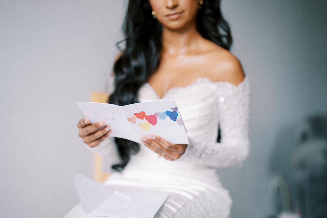 Young woman in an elegant white wedding dress sitting and holding a colorful card with hearts, capturing a tender pre-wedding moment.