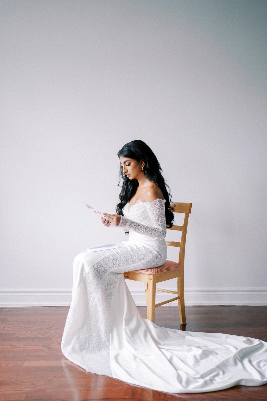 A bride in an elegant off-shoulder white wedding dress sits on a chair reading a letter, her long dark hair cascading down her shoulders, against a simple, neutral background.