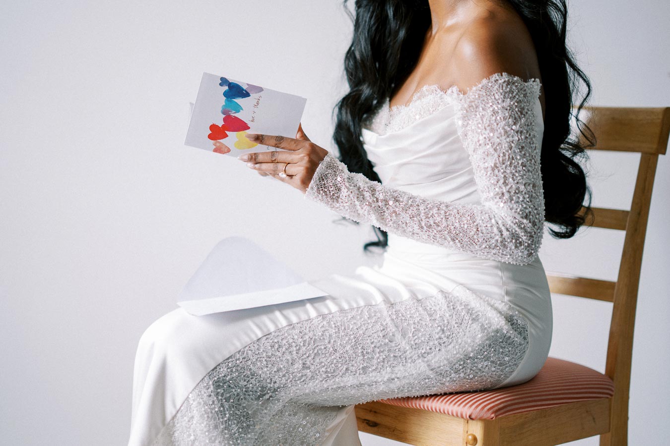 Elegant bride in a lace wedding dress reading a colorful card while seated on a wooden chair.