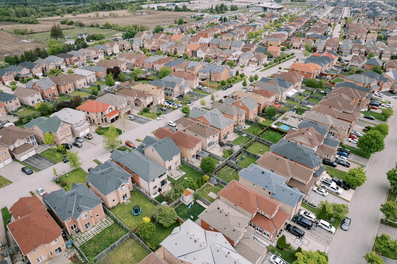Aerial view of a suburban neighborhood with rows of modern houses, varied roof colors, and green lawns linked by streets and sidewalks, surrounded by trees and open fields.