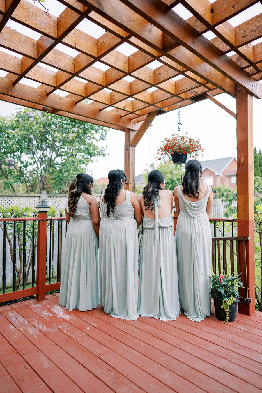 Four bridesmaids in matching light blue dresses stand on a wooden deck, beneath a wooden pergola with a hanging flower basket. Lush green trees and a fenced backyard can be seen in the background.