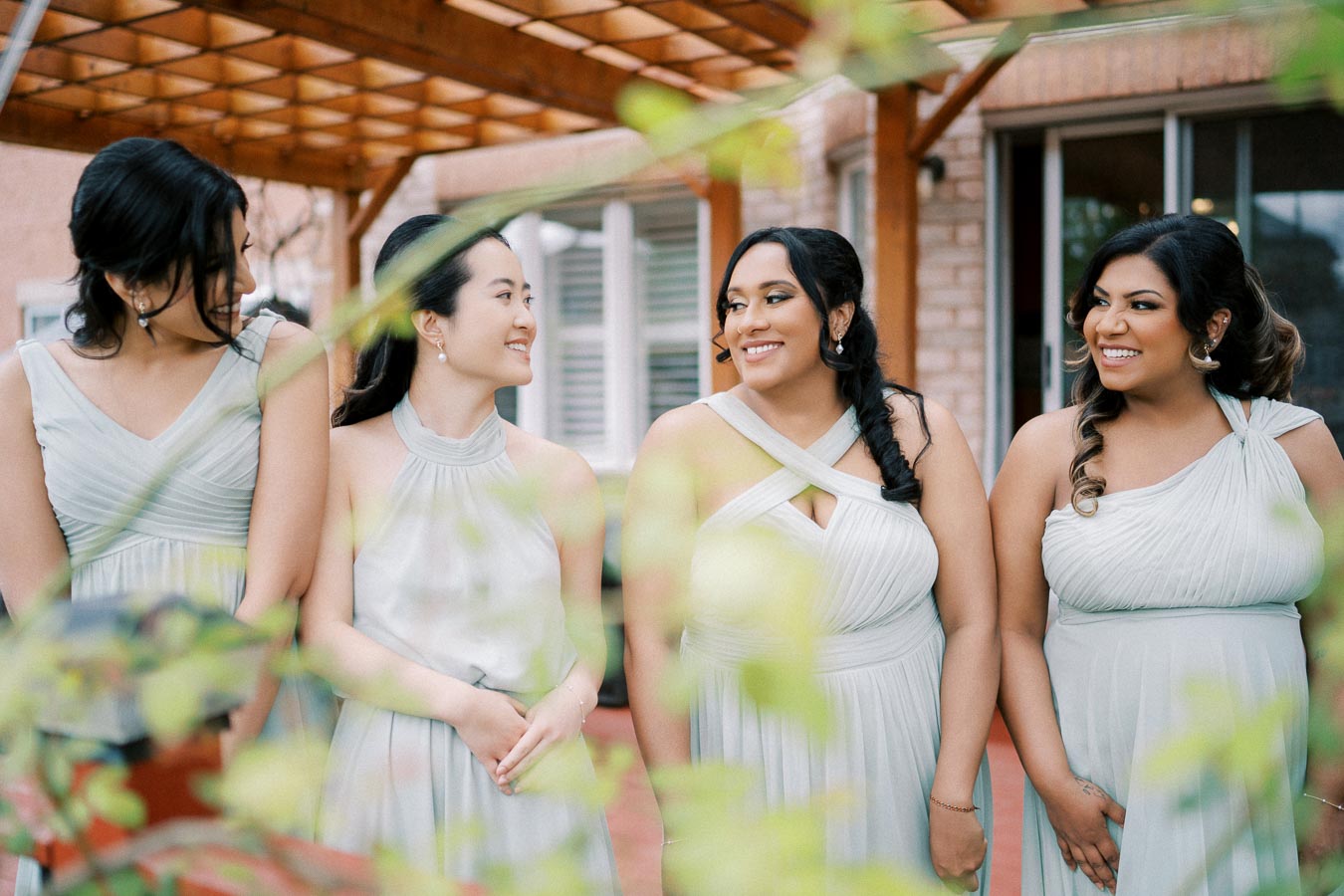 Four bridesmaids smiling and wearing elegant, matching light gray dresses while standing outside under a wooden pergola.