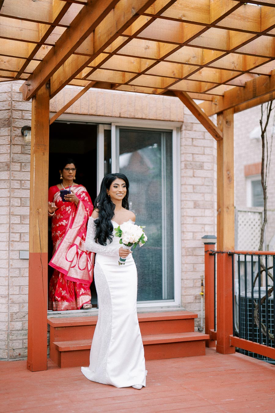 A bride in a white wedding gown holds a bouquet as she walks under a wooden pergola, with a woman in a red saree following her from a house.