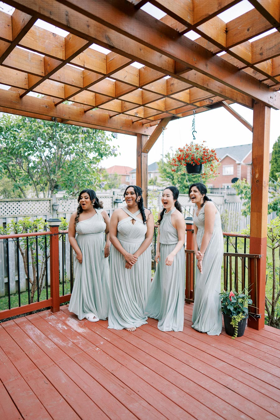 Four bridesmaids in matching light gray dresses stand on a wooden deck under a pergola, surrounded by greenery and potted plants, sharing a joyful moment together.