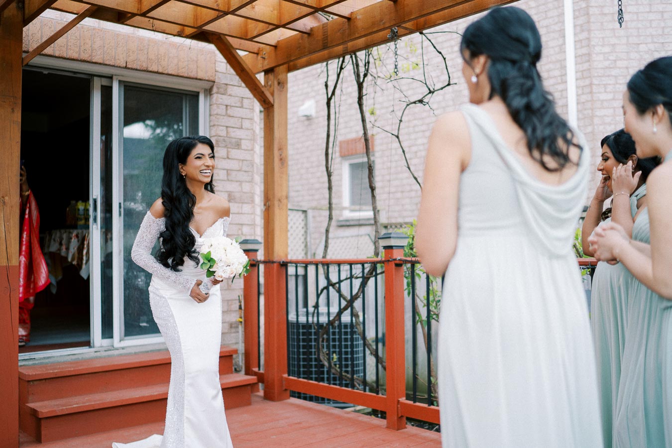 Bridal moment under pergola: A bride in a white off-the-shoulder dress and holding a bouquet smiles joyfully at bridesmaids in pastel dresses, enjoying a heartfelt moment on a wooden deck.