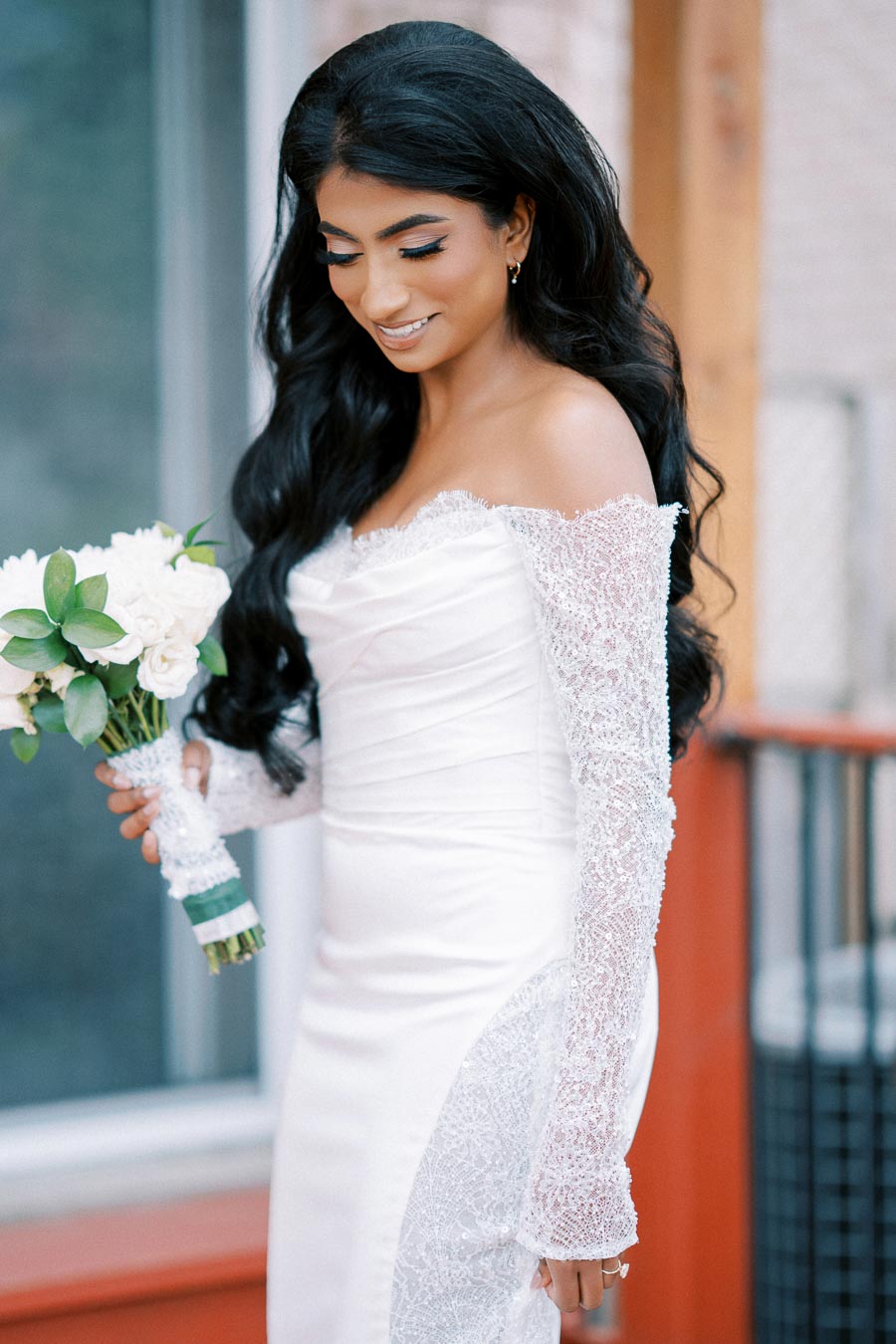 A bride in a white lace wedding dress holds a bouquet of white flowers, smiling and looking down. The image captures the elegance and joy of a wedding day.