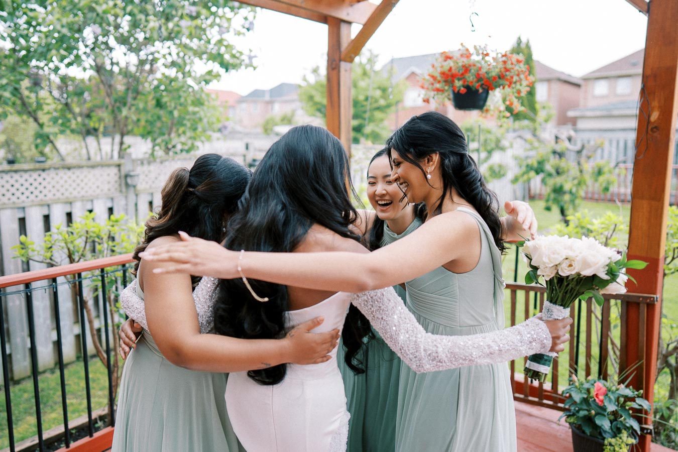 Bridal party hugging in garden setting; bride in white dress holding bouquet of white roses, surrounded by bridesmaids in mint green dresses, sharing a joyful moment before the wedding ceremony.