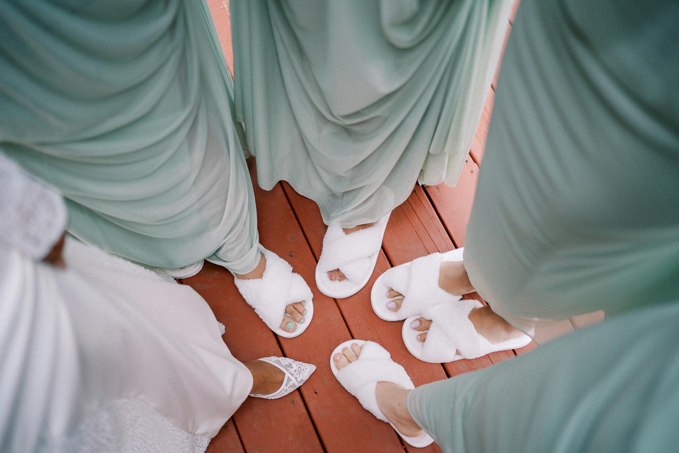 Bridesmaids wearing light green dresses and white fluffy slippers standing on a wooden deck.