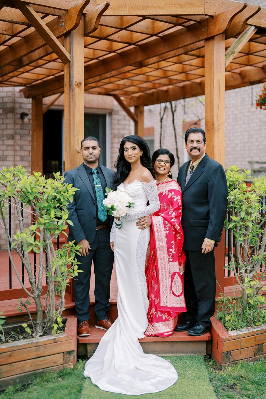 A bride in a white dress holding a bouquet stands with three family members under a wooden pergola. The family is smiling against the backdrop of a garden setting.