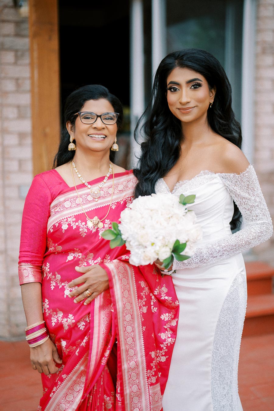 Elegant bride in white gown holding bouquet, standing beside a woman in a bright pink saree, smiling and posing outdoors on a wedding day.