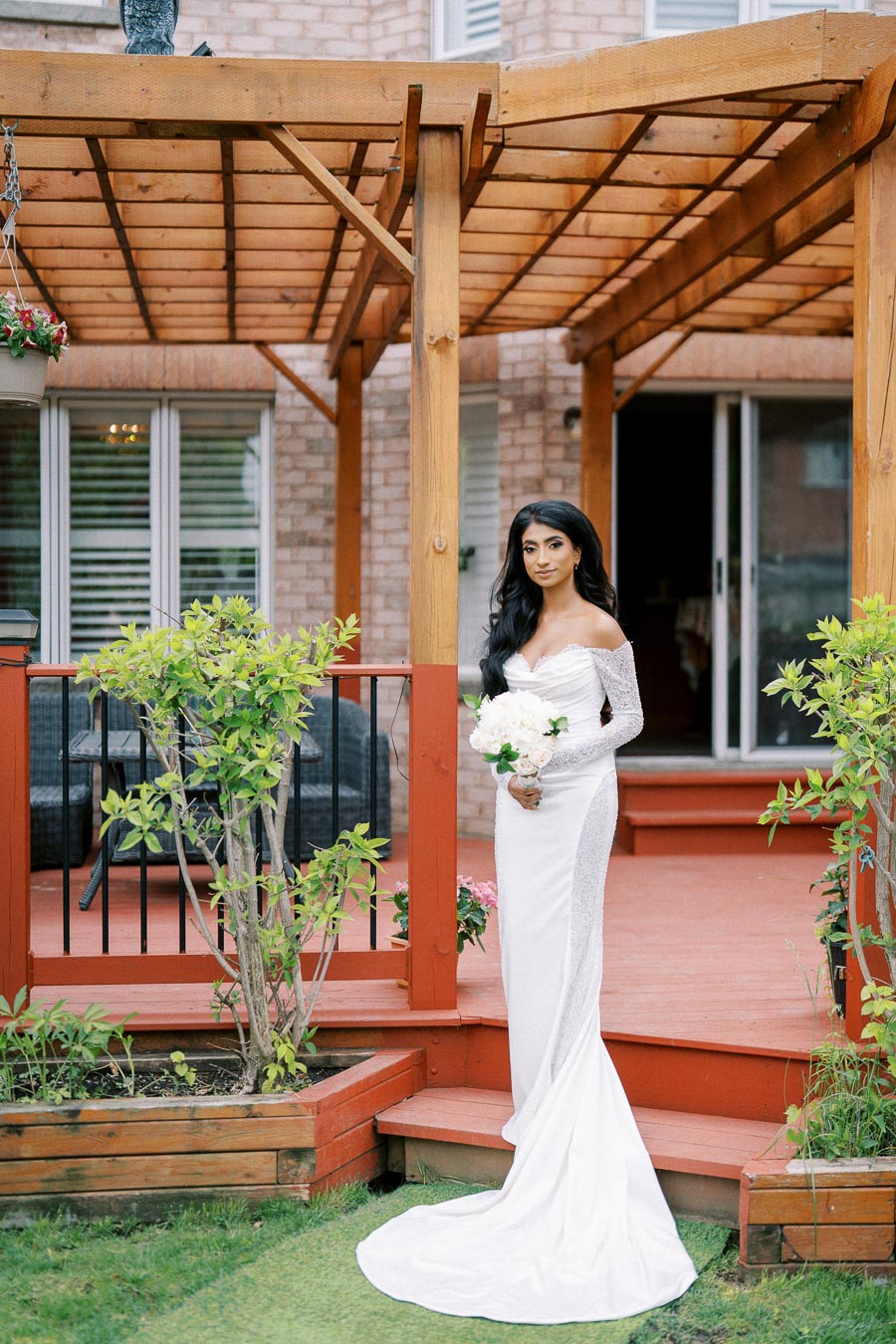 Bride in a long white dress holding a bouquet of white flowers, standing on a wooden deck with a pergola and garden plants in the background.