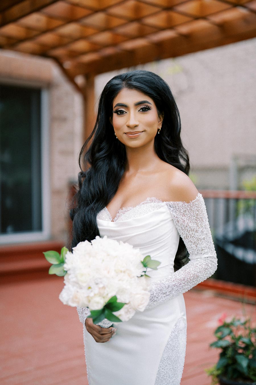 A bride in a beautiful white wedding dress with lace sleeves holds a bouquet of white flowers while standing on a wooden terrace with a trellis backdrop.