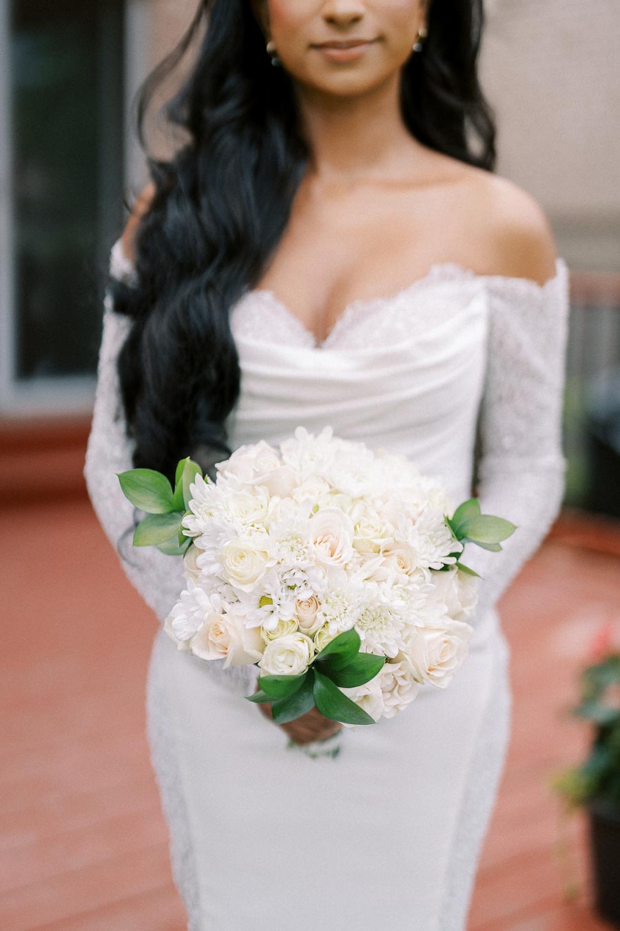 A bride in an elegant, off-the-shoulder lace wedding dress holds a bouquet of white and cream roses with greenery.