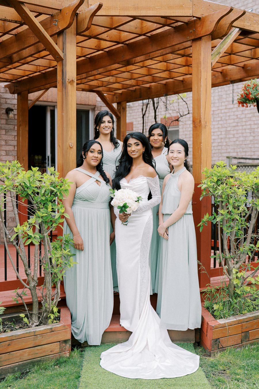 A bride in a white gown holding a bouquet poses with bridesmaids dressed in light green dresses under a wooden pergola, set against a lush garden backdrop.
