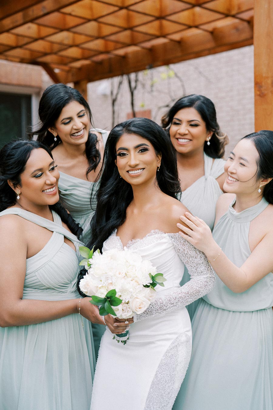 A bride in a white lace wedding dress smiling with her bridesmaids in matching light green dresses, holding a bouquet of white flowers, set in an outdoor venue with wooden pergola.