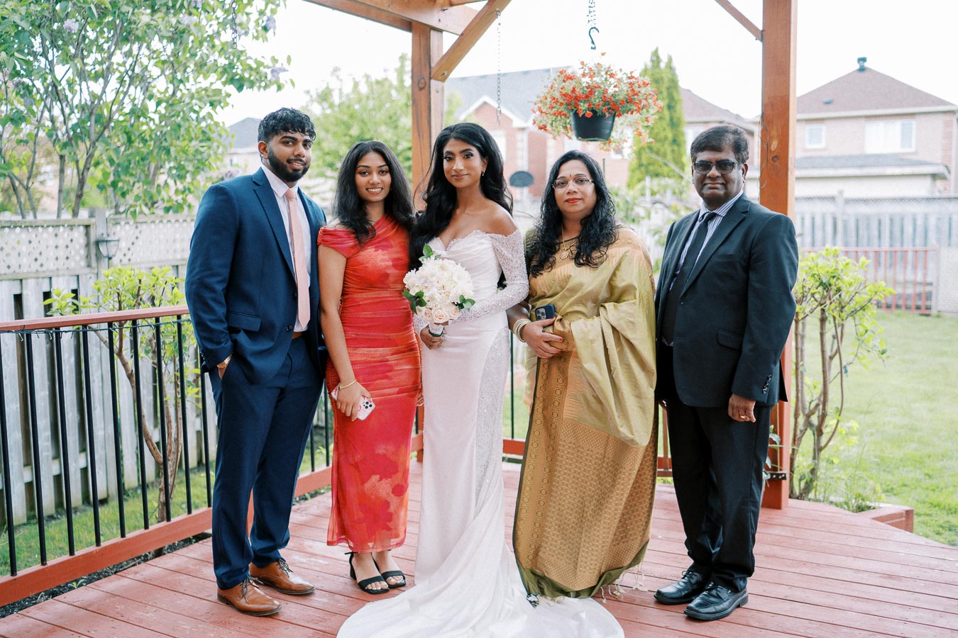 A group photo at an outdoor wedding ceremony featuring a bride in a white gown holding a bouquet, surrounded by elegantly dressed family members in traditional and formal attire, standing on a wooden deck with greenery in the background.