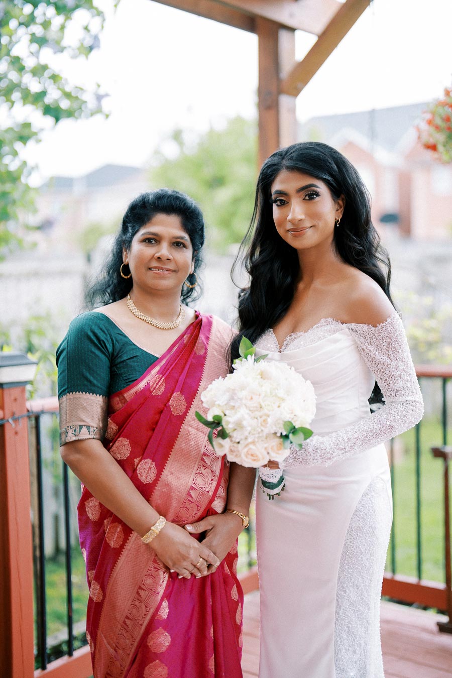 A bride in an elegant white wedding dress stands next to a woman in a vibrant pink and gold sari. They are outdoors on a wooden terrace with lush greenery in the background. The bride holds a bouquet of white flowers, and both women are smiling.