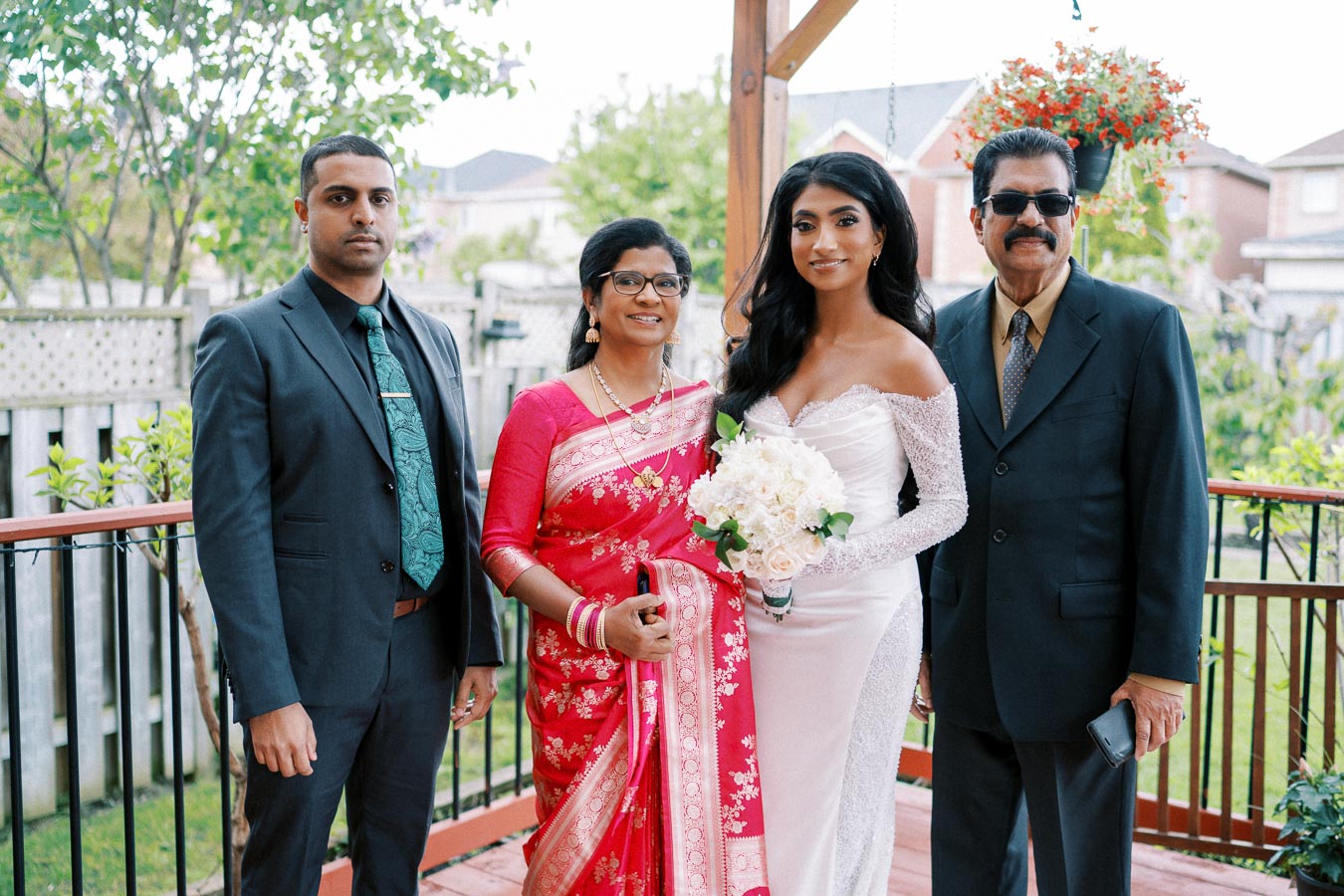 A family celebrating a wedding outdoors, featuring a bride in a white dress holding a bouquet, alongside family members in formal attire and a traditional red saree, with a garden backdrop.