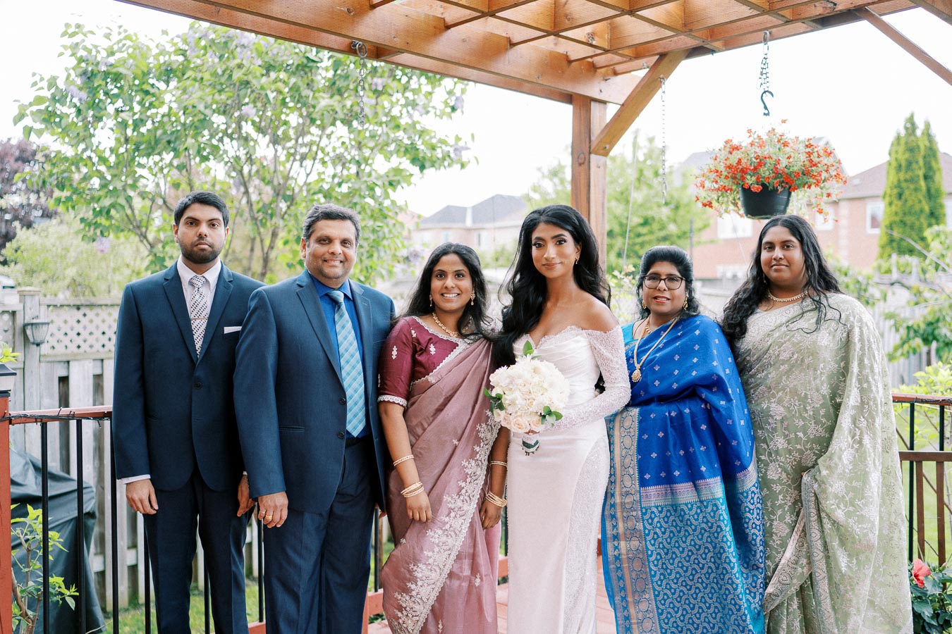 A family event with six individuals posing under a wooden pergola in a garden setting, with the central figure in a white gown holding a bouquet, surrounded by others in formal and traditional attire.