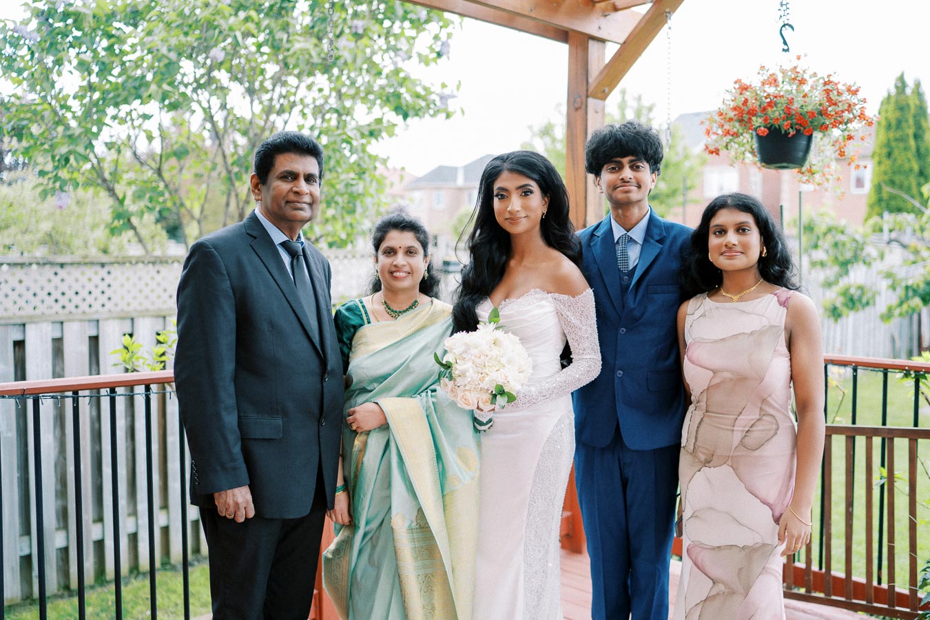 A family posing together outdoors, with a bride in a white dress holding a bouquet, surrounded by family members in formal attire, under a wooden pergola with greenery in the background.