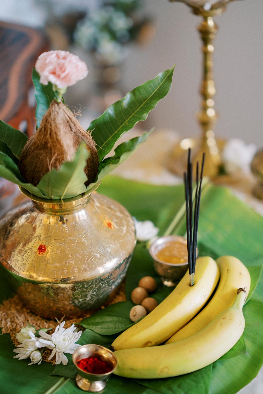 Traditional Hindu offering setup featuring a decorated coconut in a brass Kalash, bananas, incense sticks, and flower petals on a green leaf base.