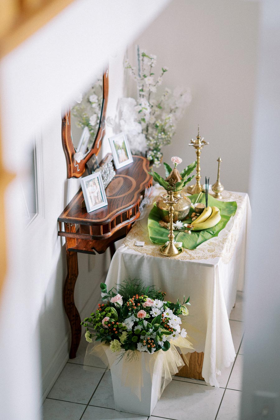 Elegant home altar with traditional decor, featuring brass lamps and a small table adorned with bananas and flowers, beside a mirror and framed photographs. A bouquet of fresh flowers adds a touch of color to the ritual setting.