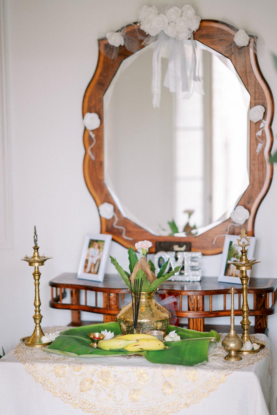 Traditional Indian offering arrangement with a decorated mirror, featuring brass lamps, bananas, a coconut, and incense sticks on a green leaf, atop an embroidered white cloth.