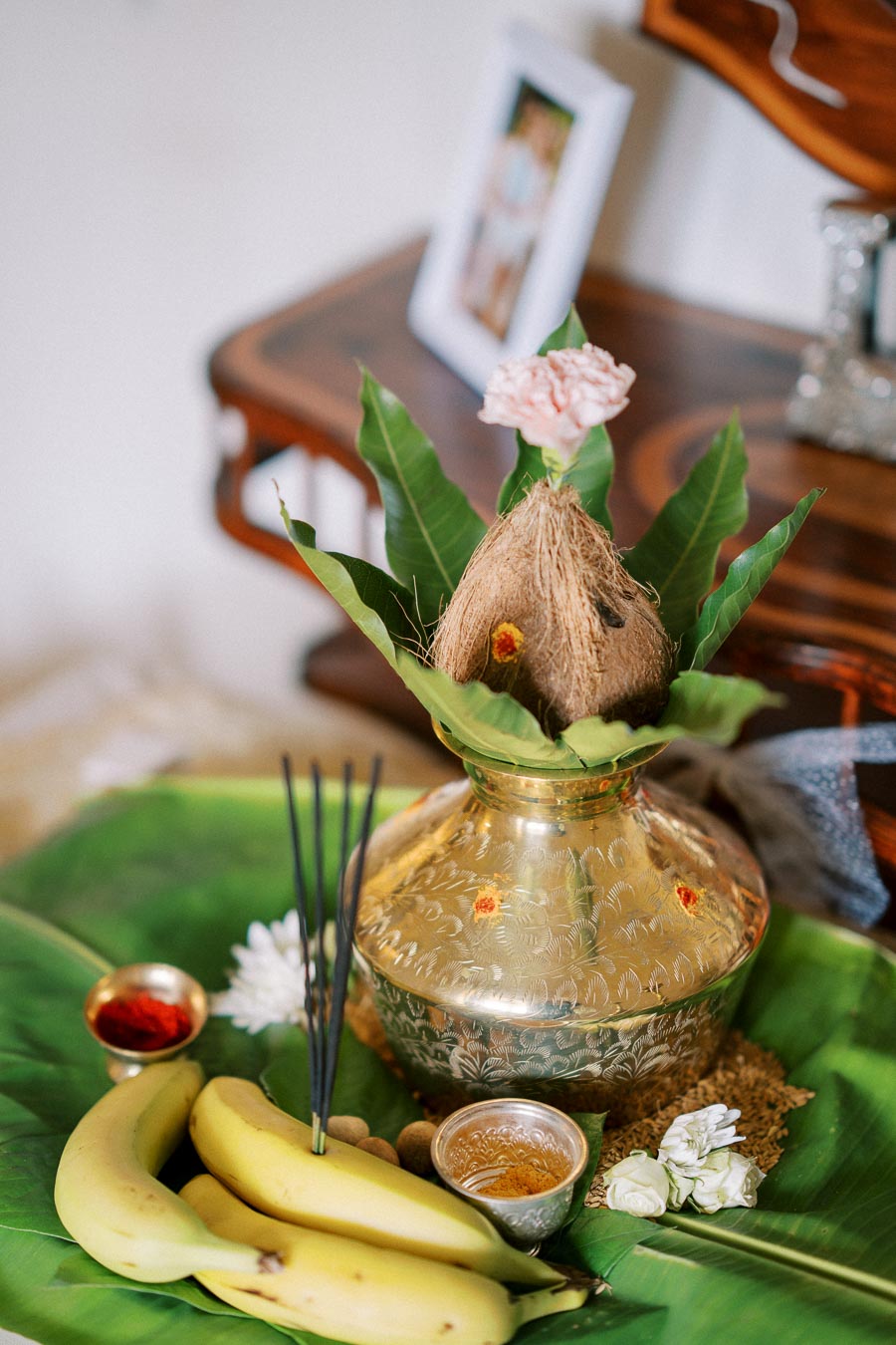 A traditional Indian puja setup with a decorative kalasha (metal pot), topped with a coconut and surrounded by mango leaves. The arrangement includes bananas, small bowls of turmeric and vermillion, and burning incense sticks, all placed on a banana leaf.