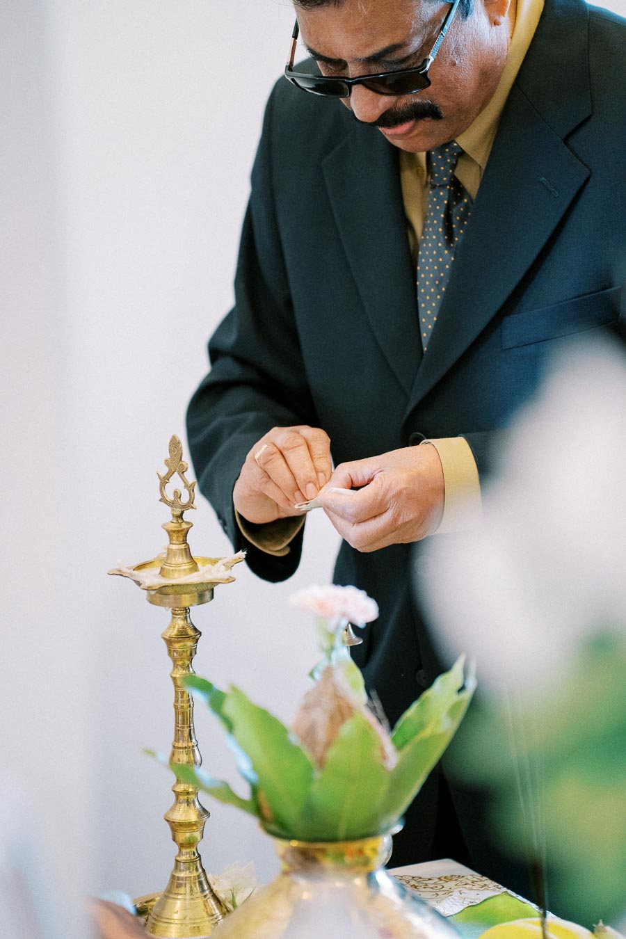 A man in a suit and tie, wearing glasses, is carefully lighting a traditional brass oil lamp in a ceremonial setting, with flowers and greenery in the foreground.