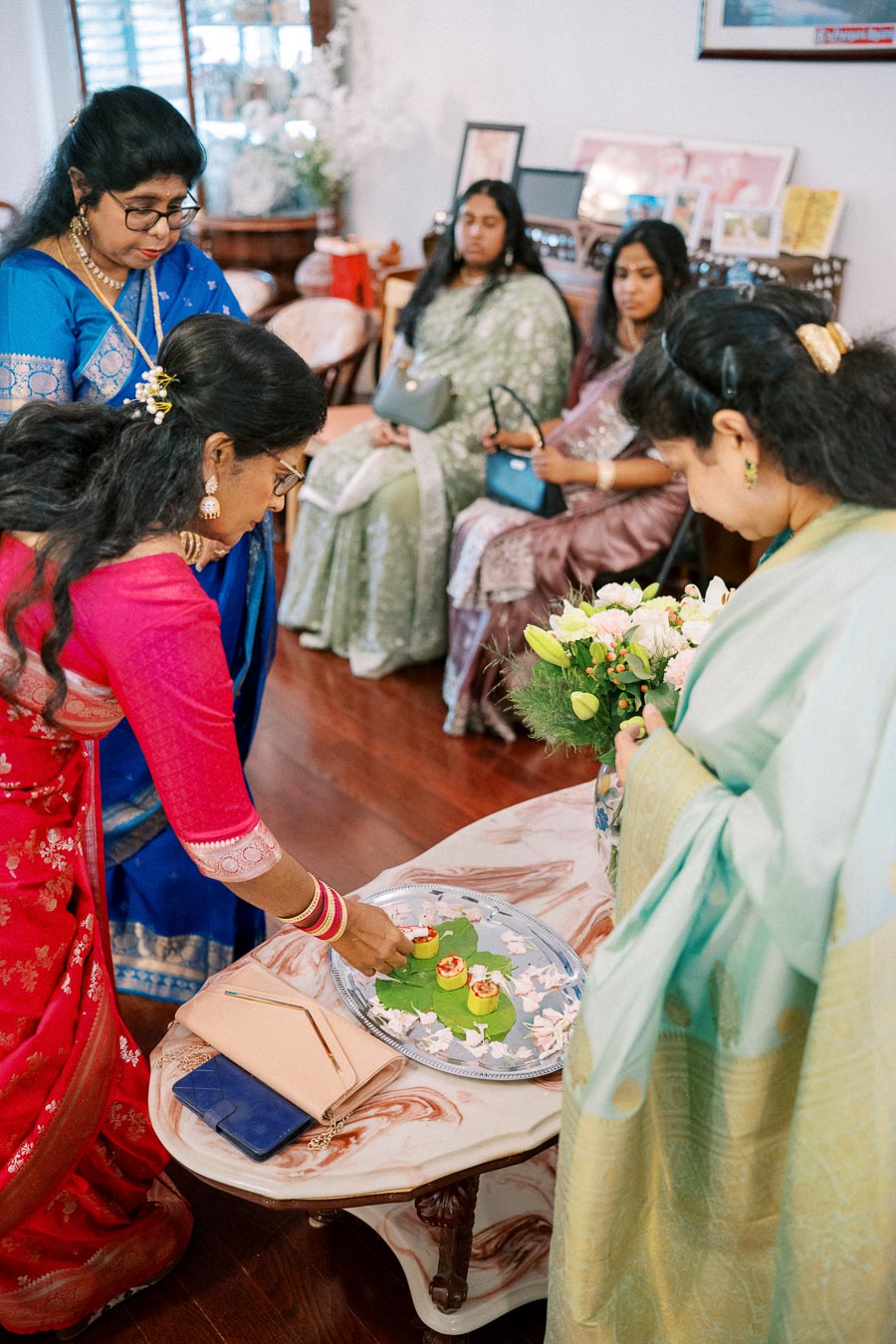 Women in colorful traditional attire partaking in a cultural ceremony, arranging items on a decorated platter in a home setting, with others seated in the background watching.