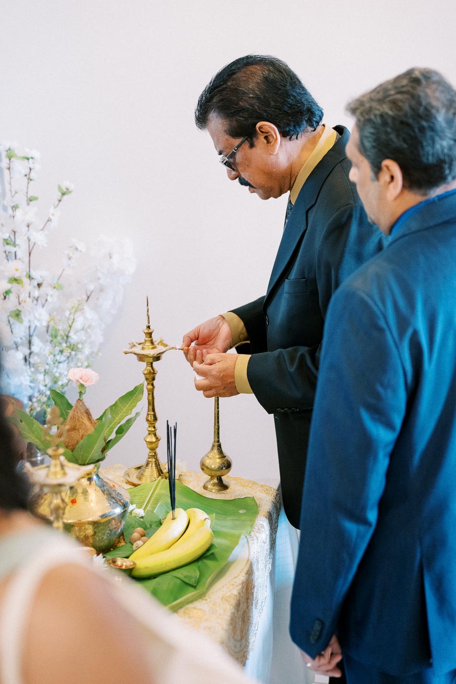Two men in suits participating in a traditional Hindu prayer ceremony, lighting a brass oil lamp on a table adorned with fresh flowers, bananas, and incense sticks, symbolizing spirituality and devotion.